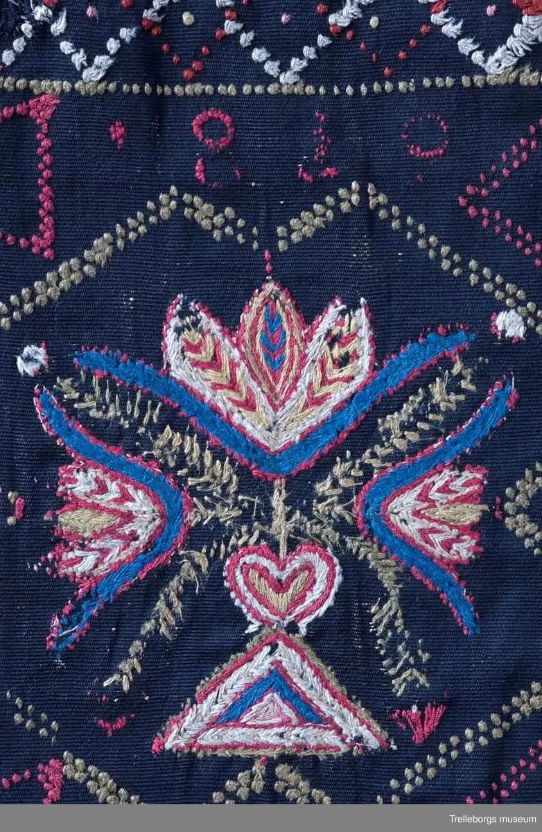 Material: varp 2 tr lingarn, inslag 1 tr ullgarn, ripsbindning. Broderi och flossaknutar 1 tr ullgarn. 
Bården runtom är vävd i tränsaflossa, diagonal inrutning. Dessutom är vävt konturer till tre rutor, dessa delar in agedynans mittfält i tre avdelningar. I tränsaflossan är även invävt konturerna till en hjärtfigur i vardera hörnet, samt två timglas utmed långsidorna. I var och en av de tre rutorna är broderat en blomstervas med tre tulpaner. Färgerna är här blått, cyklamen, gult, vitt och olivgrönt.

Tidigare konserverad då bitarna har sytts ihop på ny svart botten. Eventuellt kan det vara så att dynan klippts sönder för att brukas i två delar eller vid arvsskifte.
