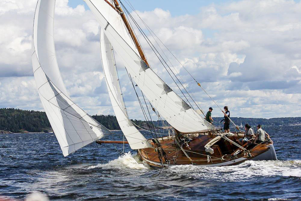 Skøyta S/Y Venus i vannet, tre seil, fire personer ombord. I bakgrunnen utsikt til skog.