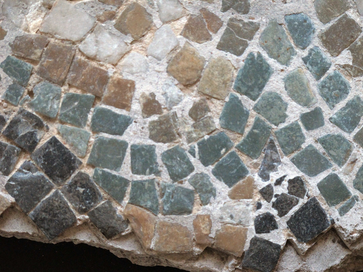 Mangefarget mosaikk fragment, tilnærmet rektangulært av form. Grønne, hvite, grå og gule tesseraer i naturstein, satt i fin kornet, hvit mørtel. Steinene av ulik størrelse, satt bueformede mønster.