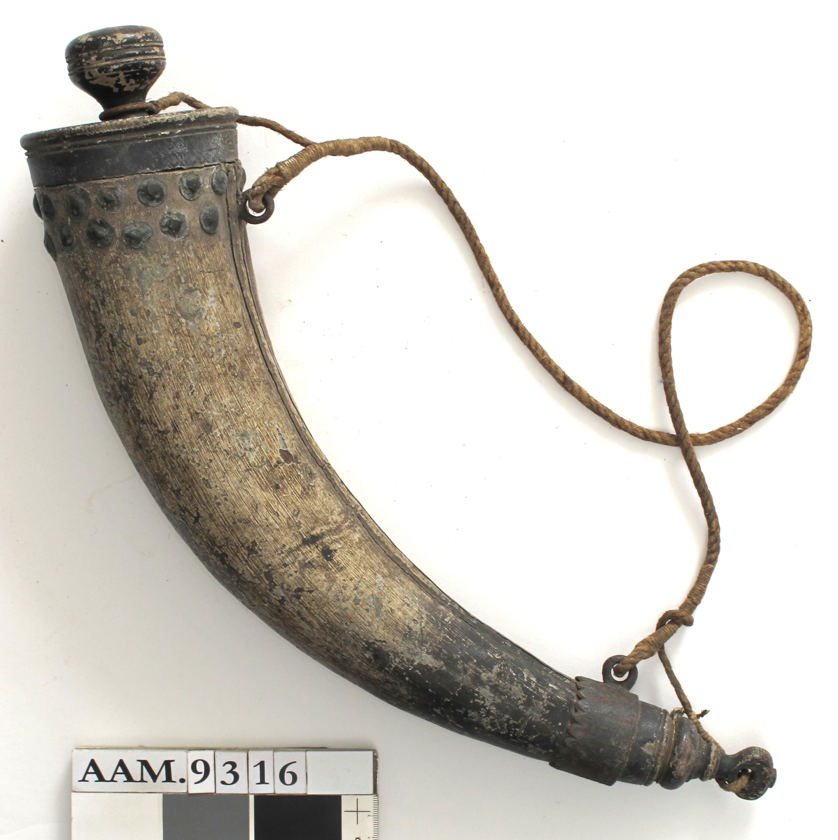 Krutthorn, imitert horn av malt tre,  med sortmalt trelokk og bunn, jernbeslag og   messingknapper. Horn malt i hornimitasjon med lysbrune striper, sort i nedre del.  Skjøt langs innsiden av buen. Sort  dreiet  lokk med nedsenket toppflate, stor dreiet knott med skrugjenger.  Under trelokket to rader med messingknapper. I nedre del et jernbeslag med takker langs overkanten, i dette en jernring  for feste av bæresnoren av tau.