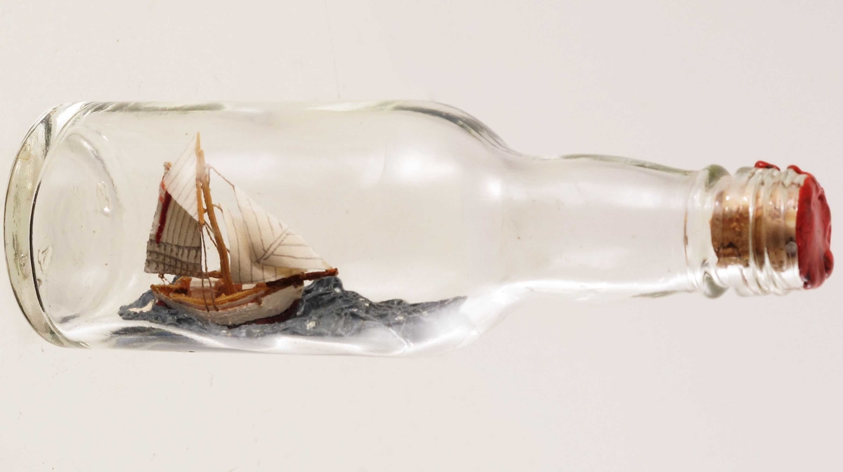 Mini flaskeskip: Losbåt.  Glass, klart. Kork med rød lakk. Hvitmalt miniatyrskute med rød bunn og rød stripe i seilet. Blå sjø.