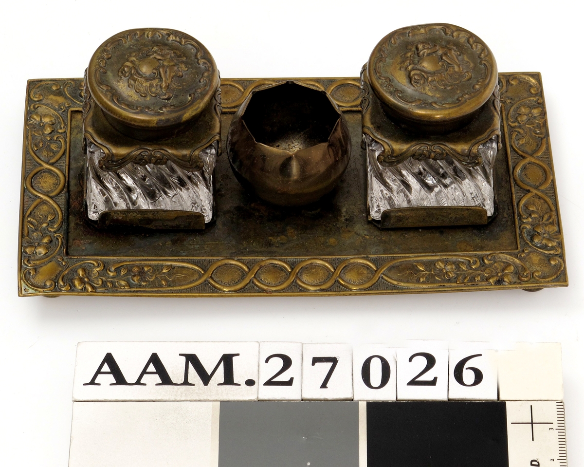 Skrivestell. Presset  messing, 2 blekkhus av  pressglass. Rektangulær messingplate med presset ornamentbord langs kanten og 4 små dreide føtter. Presset dekorativt mønster.
