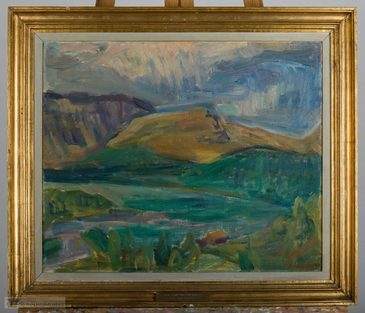 Fjellandskap. Et fjellvann innrammet av vegetasjon med fjell og heier i bakgrunnen.