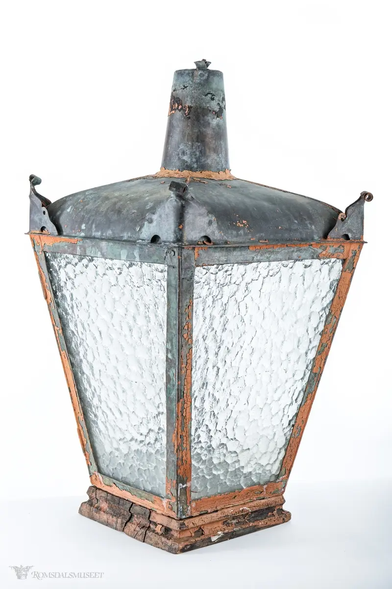 Rødmalt parafingatelykt i metall med spraglete glass og pipe. Den ene siden er hengslet og kan låses med en en gjennomgående metallring med krok. Lykten er montert på en trebunn satt sammen av tre tykke profilerte bord. Innvendig er det boret fem hull, et i midten og et på hver av sidene, for festing av brenner. Hvert hjørne av taket på lykten har ornamenteringer. Brukt i Molde.