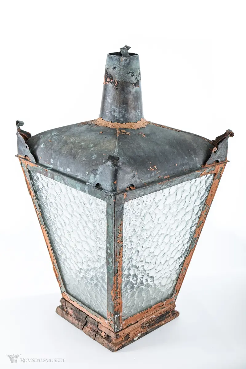 Rødmalt parafingatelykt i metall med spraglete glass og pipe. Den ene siden er hengslet og kan låses med en en gjennomgående metallring med krok. Lykten er montert på en trebunn satt sammen av tre tykke profilerte bord. Innvendig er det boret fem hull, et i midten og et på hver av sidene, for festing av brenner. Hvert hjørne av taket på lykten har ornamenteringer. Brukt i Molde.