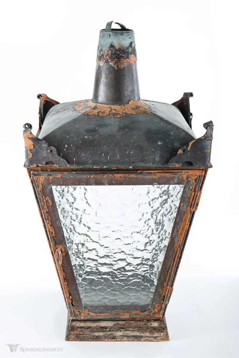 Rødmalt parafingatelykt i metall med spraglete glass og pipe. Den ene siden er hengslet og kan låses med en en gjennomgående metallring med krok. Lykten er montert på en trebunn satt sammen av tre tykke profilerte bord. Innvendig er det boret fem hull, et i midten og et på hver av sidene, for festing av brenner. Hvert hjørne av taket på lykten har ornamenteringer. Brukt i Molde.