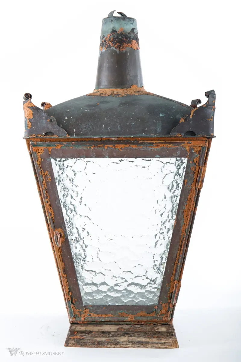 Rødmalt parafingatelykt i metall med spraglete glass og pipe. Den ene siden er hengslet og kan låses med en en gjennomgående metallring med krok. Lykten er montert på en trebunn satt sammen av tre tykke profilerte bord. Innvendig er det boret fem hull, et i midten og et på hver av sidene, for festing av brenner. Hvert hjørne av taket på lykten har ornamenteringer. Brukt i Molde.
