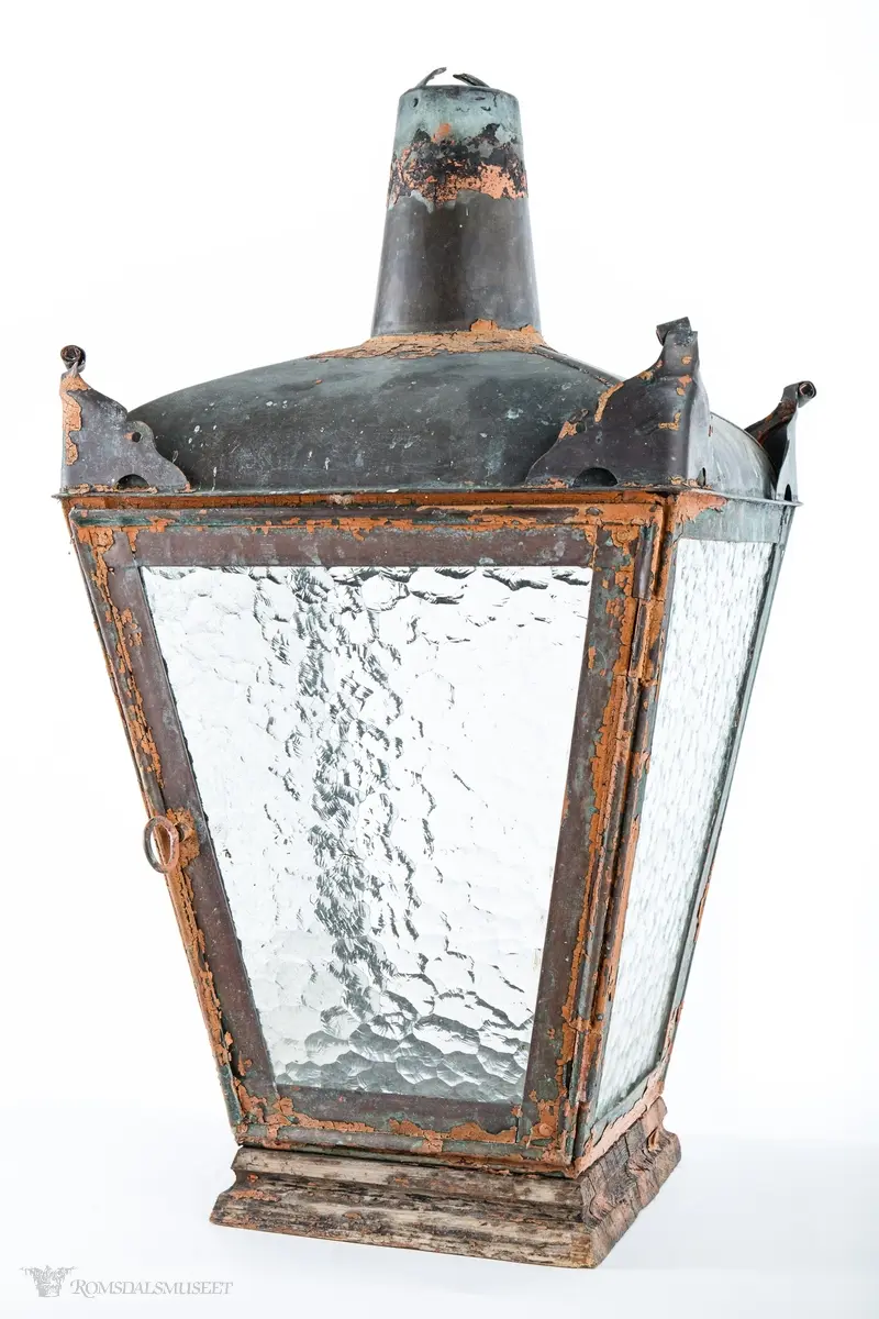 Rødmalt parafingatelykt i metall med spraglete glass og pipe. Den ene siden er hengslet og kan låses med en en gjennomgående metallring med krok. Lykten er montert på en trebunn satt sammen av tre tykke profilerte bord. Innvendig er det boret fem hull, et i midten og et på hver av sidene, for festing av brenner. Hvert hjørne av taket på lykten har ornamenteringer. Brukt i Molde.