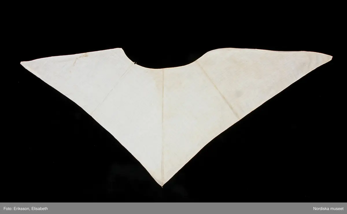 Huvudliggaren:
"Sorgdräkt för kvinna. Särk med 'hängseltåg', 'särkyvdel'; vit -'häsklä'; blått livstycke; svart tröja; huvudduk; 'svart näsduk'; hatt; 'raskförklä', 'sockar',  'hårbandsvifsa', huvudbindel."

Bilaga:
a: "8 Särk, övdel. av linne eller blågarn. med broderade linningar på överdelen"
c: "11. Hvit häsklä."
Anm: Kung Kerstin Larsdotters resa i Floda sn Dalarna 24/2 - 8/3 1912. Innehåller förteckning på inköp av dräktdelar från Floda, inv nr 117 216 - 117 239. Här finns  tillverkningstid och beskrivningar på vissa av dräktdelarna.  I vissa fall nämns också utförligt på vilka helgdagar dräktdelen  användes.

Katalogkort:
a: Hängselsärk
b. Överdel av halvlinne vävt i kypert.
c.  Halskäde av småmönstrat bomullstyg, liknande kambris, vävt  i kypertvariation med drällmönster.
d. Livstycke av mörkblå kraftig vadmal,
e. Tröja av grov svart vadmal

i. 
 Mönstervävt band med upplockat mönster i rött z-tvinnat ullgarn på vit linnebotten, med hjärtan, kryss och rombformer, 2 band om vardera 124 cm har vikts och sytts ihop så att det ena bildar en ögla och det andra har fria ändar avslutade med en tofs i bandets garn.
Läs mer om dräktskicket i Floda i: Albert Alm, Dräktalmanacka för Floda socken, Nordiska museet, Sthlm 1930
/Berit Eldvik 2005-04-14