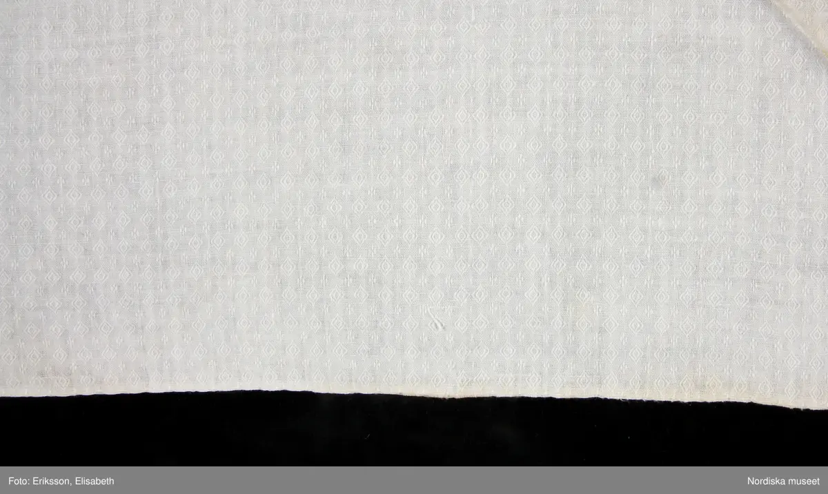 Huvudliggaren:
"Sorgdräkt för kvinna. Särk med 'hängseltåg', 'särkyvdel'; vit -'häsklä'; blått livstycke; svart tröja; huvudduk; 'svart näsduk'; hatt; 'raskförklä', 'sockar',  'hårbandsvifsa', huvudbindel."

Bilaga:
a: "8 Särk, övdel. av linne eller blågarn. med broderade linningar på överdelen"
c: "11. Hvit häsklä."
Anm: Kung Kerstin Larsdotters resa i Floda sn Dalarna 24/2 - 8/3 1912. Innehåller förteckning på inköp av dräktdelar från Floda, inv nr 117 216 - 117 239. Här finns  tillverkningstid och beskrivningar på vissa av dräktdelarna.  I vissa fall nämns också utförligt på vilka helgdagar dräktdelen  användes.

Katalogkort:
a: Hängselsärk
b. Överdel av halvlinne vävt i kypert.
c.  Halskäde av småmönstrat bomullstyg, liknande kambris, vävt  i kypertvariation med drällmönster.
d. Livstycke av mörkblå kraftig vadmal,
e. Tröja av grov svart vadmal

i. 
 Mönstervävt band med upplockat mönster i rött z-tvinnat ullgarn på vit linnebotten, med hjärtan, kryss och rombformer, 2 band om vardera 124 cm har vikts och sytts ihop så att det ena bildar en ögla och det andra har fria ändar avslutade med en tofs i bandets garn.
Läs mer om dräktskicket i Floda i: Albert Alm, Dräktalmanacka för Floda socken, Nordiska museet, Sthlm 1930
/Berit Eldvik 2005-04-14