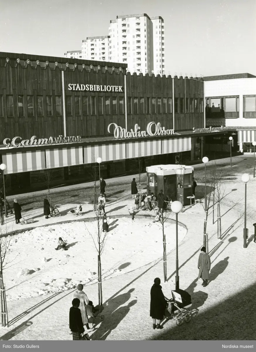 Vy över Farsta centrum med Stadsbiblioteket, G Gahns väskor och Martin Olsson. Flanerande människor och lekande barn.