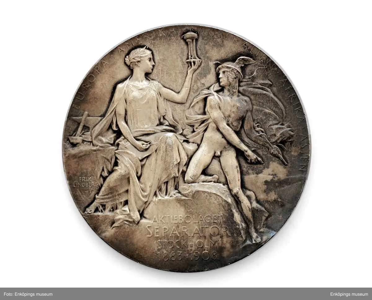 Medalj utgiven av A.B. separator, av silver. Åtsida: dubbelporträtt i högerprofil av Gustaf de Laval och JohnBernström. Frånsida: allegorisk bild med Hermes/ Merkurius med sin bevingande huvudbonad och stav samt kvinnogestalt med passare och ett okänt föremål; inskription: " EUROPA AMERIKA AUSTRALIEN AFRIKA ASIEN- AKTIEBOLAGET SEPARATOR STOCKHOLM 1883- 1908". Samt gravörens namn, Erik Lindberg.