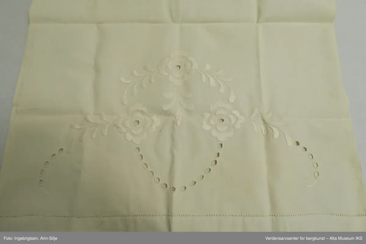 Stort pyntehåndkle med broderi i hver ende. Det ene broderiet er større enn det andre. Det store broderiet er av blomster og blader, mens det minste ser ut til å være et monogram eller lignende.