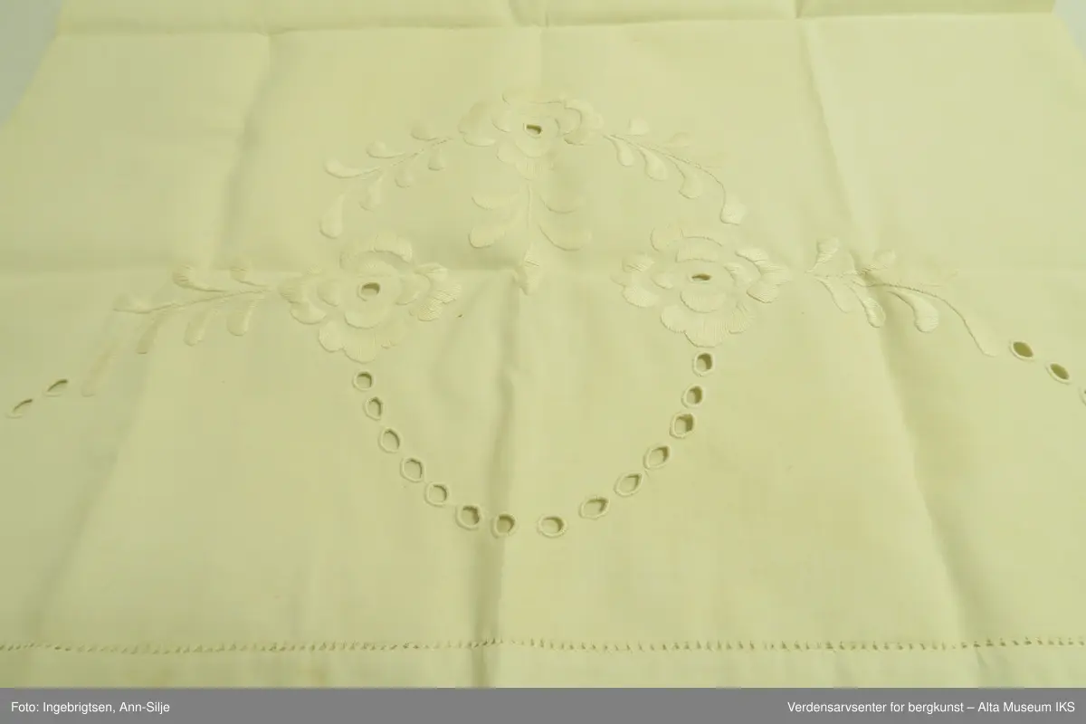 Stort pyntehåndkle med broderi i hver ende. Det ene broderiet er større enn det andre. Det store broderiet er av blomster og blader, mens det minste ser ut til å være et monogram eller lignende.