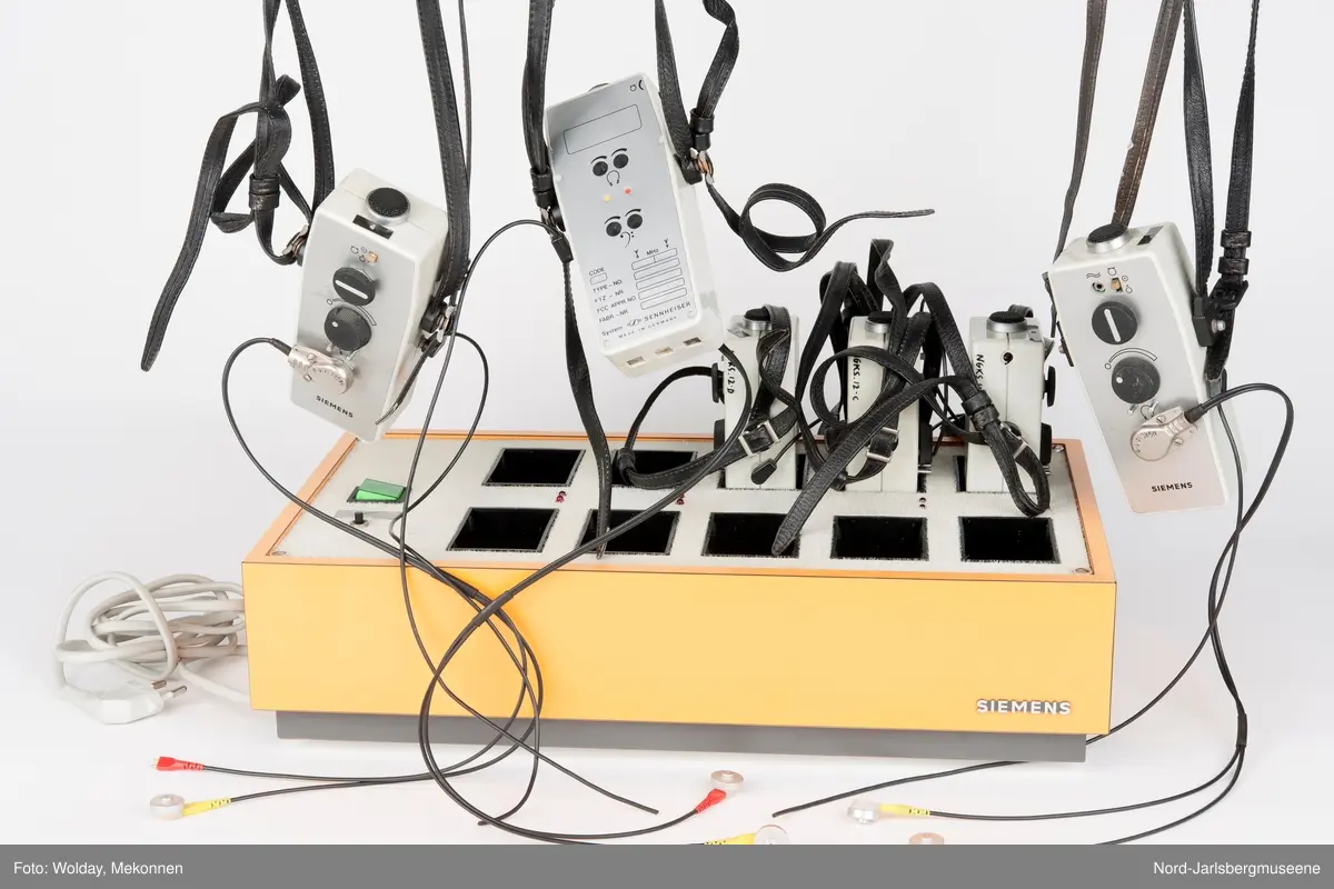 Rektangulær kasse med strømbryter og ladehull for 10 teleslynger. Kassern er oransje, telsyngene i plast og metall. Her følger det 6 teleslyngapparater med.
10 deler tilsammen