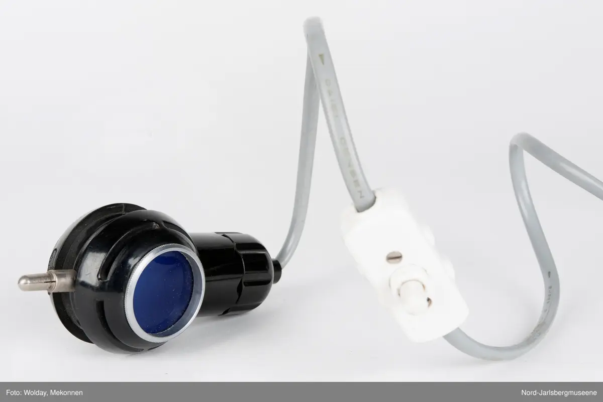 En liten lampe med to sider; en med blått lys, en med dempet lys. På toppen en metallpinne (el-kontakt?)Nyere ledning med bryter påmontert. Usikker bruksmåte og formål.