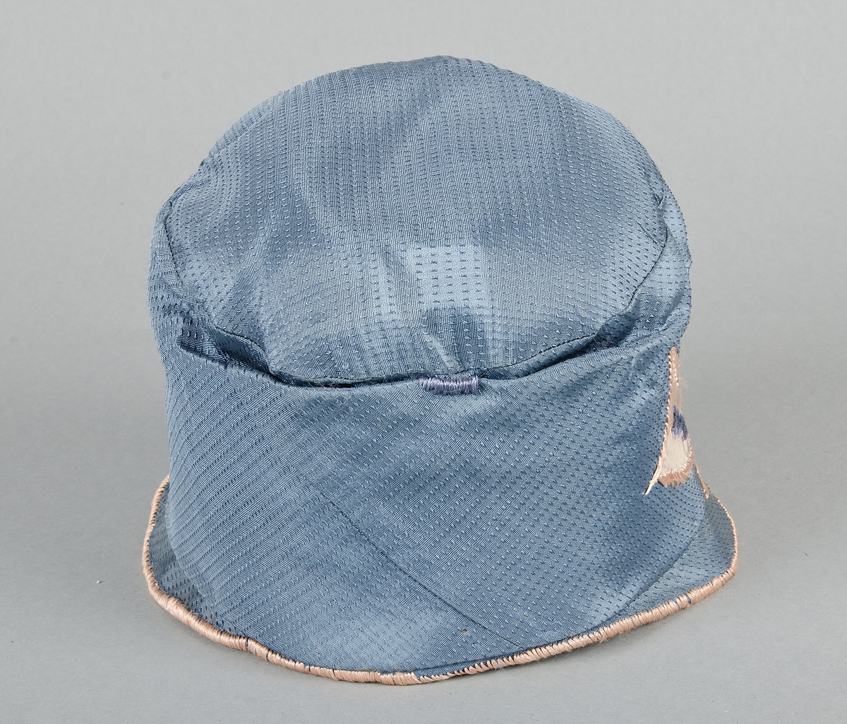 Damhatt av blått mönstrat siden med applikationer i ljusbrunt siden och broderier med blått och ljusbrunt silke. Skärmen fodrad med samma tyg som applikationerna. Fodrad med vitt tyg. Vit prislapp med svart text.