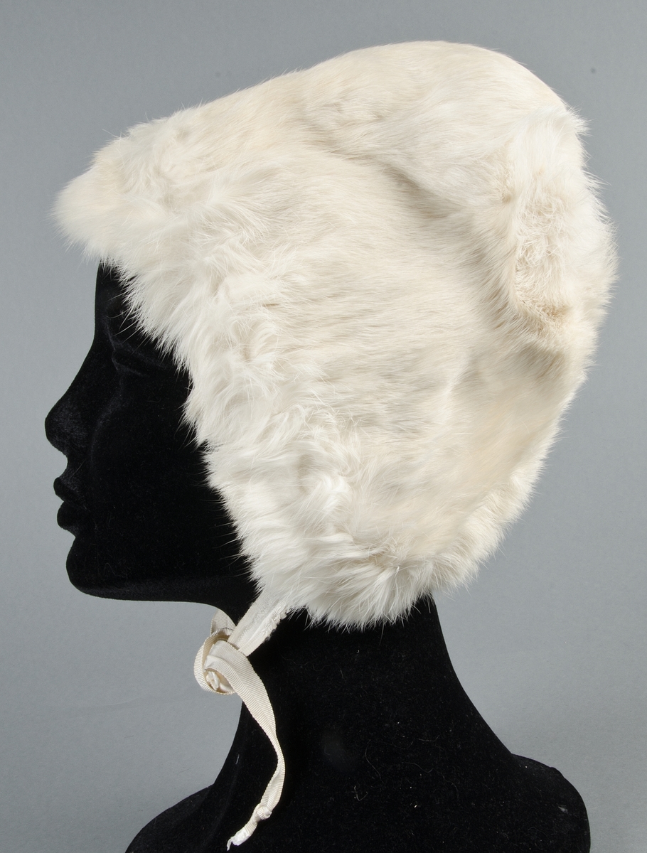 Muff med vitt kaninskinn på framsidan och grå sammet på baksidan, ficka med blixtlås på baksidan. Fodrad med vit bomullslärft, stoppad. Ingen hängband.