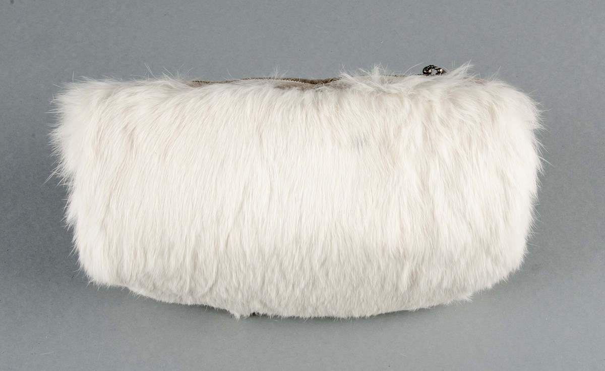 Muff med vitt kaninskinn på framsidan och grå sammet på baksidan, ficka med blixtlås på baksidan. Fodrad med vit bomullslärft, stoppad. Ingen hängband.