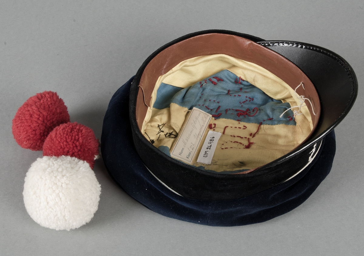 Skolmössa, från sista ring i gymnasiet, av blå sammet med svart sammetsband runt kullen och gul snodd över, skärm av svart lack. Märket med lyra och två lagerkvistar har flyttats bak på mössan och under det är fastsytt tre snoddar med en stor vit  (diam 6 cm) och två mindre (diam 5 cm) runda tofsar av ullgarn. På kullen rund vit applikation och trassel av grå tråd samt broderat med röda kedjestygn: I DIMMA DOLD, på skärmen tre broderade  vita frågetecken av kedjestygn. Invändigt fodrad med ul och blå bomullssatin, plastetikett: Marianne Adolfsson L III:3 Uppsala H.A.L. Brun svettrem.