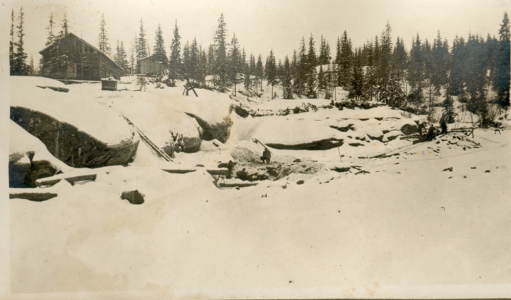 Vinterlandskap, ved elv. Snø og is. Skog. Noen bygninger. Det står noen ved elvebredden. Start av utbygging av Reinfossen kraftanlegg.