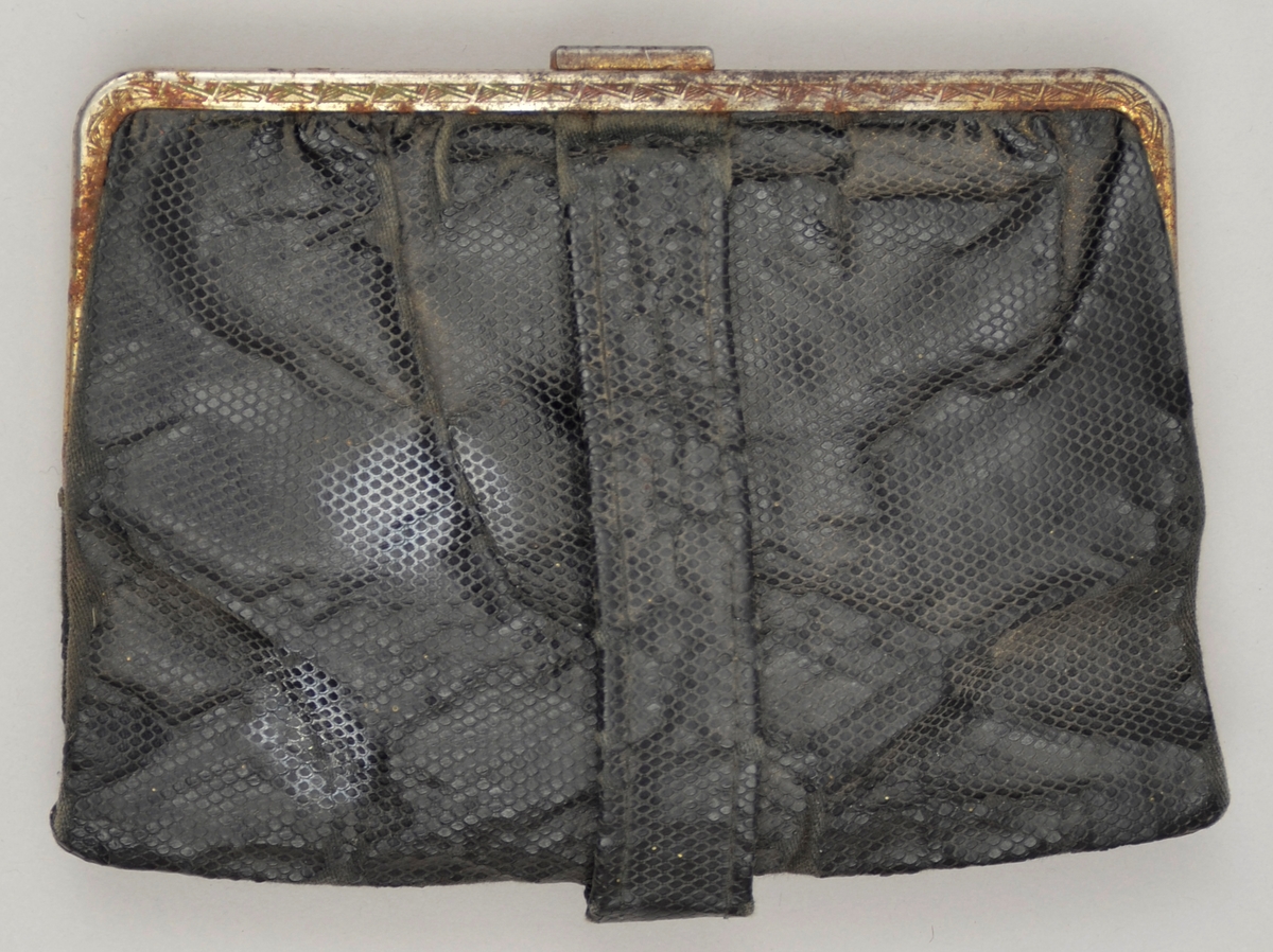 Lita handveske utan handtak. Svart skinnimitasjon. Lukking med metall-lås. Kant i metall. Lomme med knepping inni. Fôra med bomullsstoff. To kalenderlappar med dato 10 og 13 mai 1944 oppi lomma.