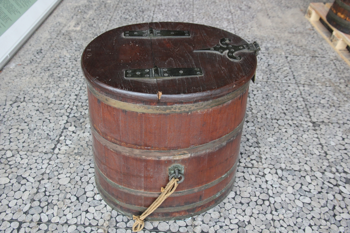 En tønne med lokk og metallhengsler. Ovale tønne med leddet lokk. Beslag og tønneband i metall. Ringer på hver side til feste av tau.