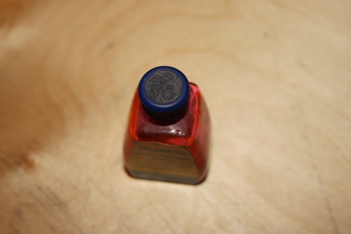 Blekkflaske med skrukork. Glassflaske med papirmerkelapp. Flasken har avrundede former/hjørner. I skrulokket er det festet en pensel.