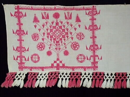 Broderat hängkläde i hallandssöm med botten i tuskaftat vitt linne. Ljusrött (rosa) broderi med tre partier och mönster. Två isydda garnhankar av lintråd i överkanten samt två små näst som inhållning. Påsydd frans i partivis röd och vit bomull med omknutna tofsar längs nederkanten. Frans +:70  mm.