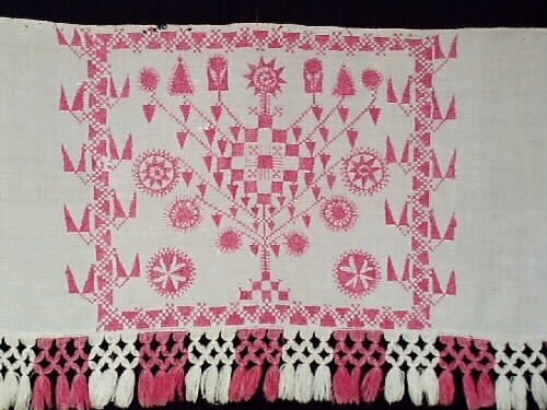 Broderat hängkläde i hallandssöm med botten i tuskaftat vitt linne. Ljusrött (rosa) broderi med tre partier och mönster. Två isydda garnhankar av lintråd i överkanten samt två små näst som inhållning. Påsydd frans i partivis röd och vit bomull med omknutna tofsar längs nederkanten. Frans +:70  mm.