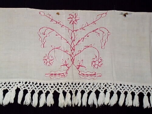 Hängkläde, del av. En liten bit av ett broderat hängkläde med hallandssöm. Bottentyg i tuskaftat vitt linne med ljusrött (rosa) broderi. Påsydd vit frans av bomull med omknutna tofsar i nederkanten. Längd= 100 cm. Bredd= 23,5 cm. Längd (frans)= 8,5 cm.