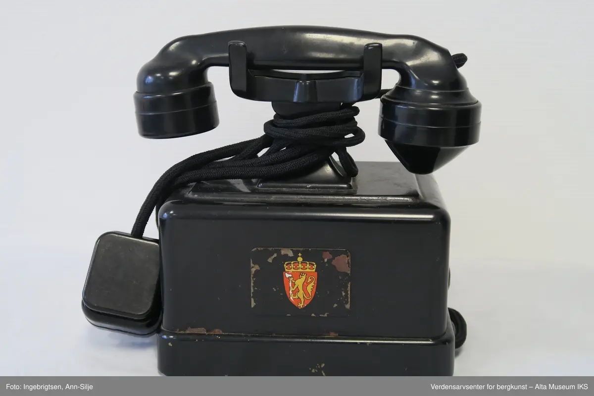 Prismeformet boks med bæreklo på toppen og telefonrør. Emblem med det norske riksvåpenet på framsiden av boksen. På siden er det festet en strømledning med støpsel i enden.