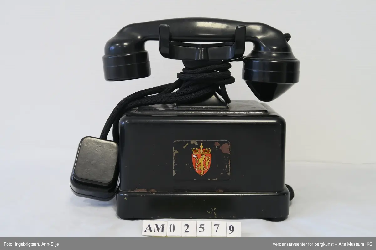 Prismeformet boks med bæreklo på toppen og telefonrør. Emblem med det norske riksvåpenet på framsiden av boksen. På siden er det festet en strømledning med støpsel i enden.