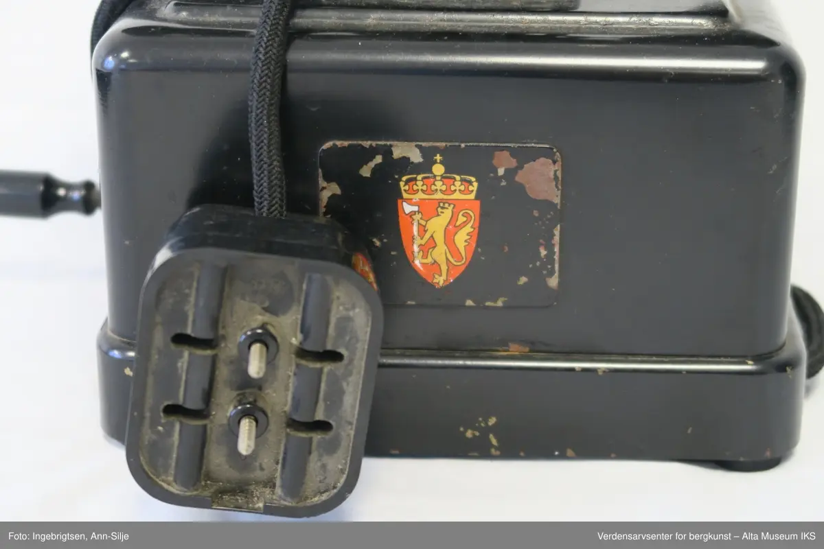 Prismeformet boks med bæreklo på toppen og telefonrør. Emblem med det norske riksvåpenet på framsiden av boksen. På siden er det festet en strømledning med støpsel i enden.