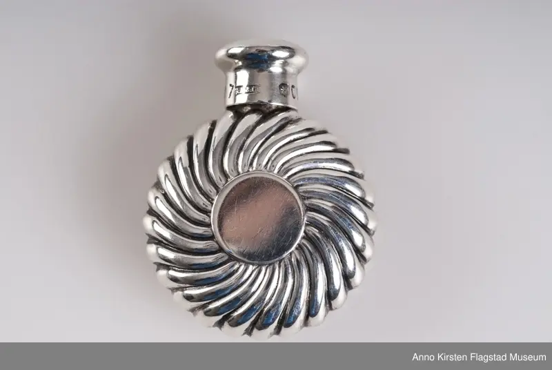 Rund. Det ene eksemplaret ligger i et sort etui med hengslet lokk (55x55x23 mm). 