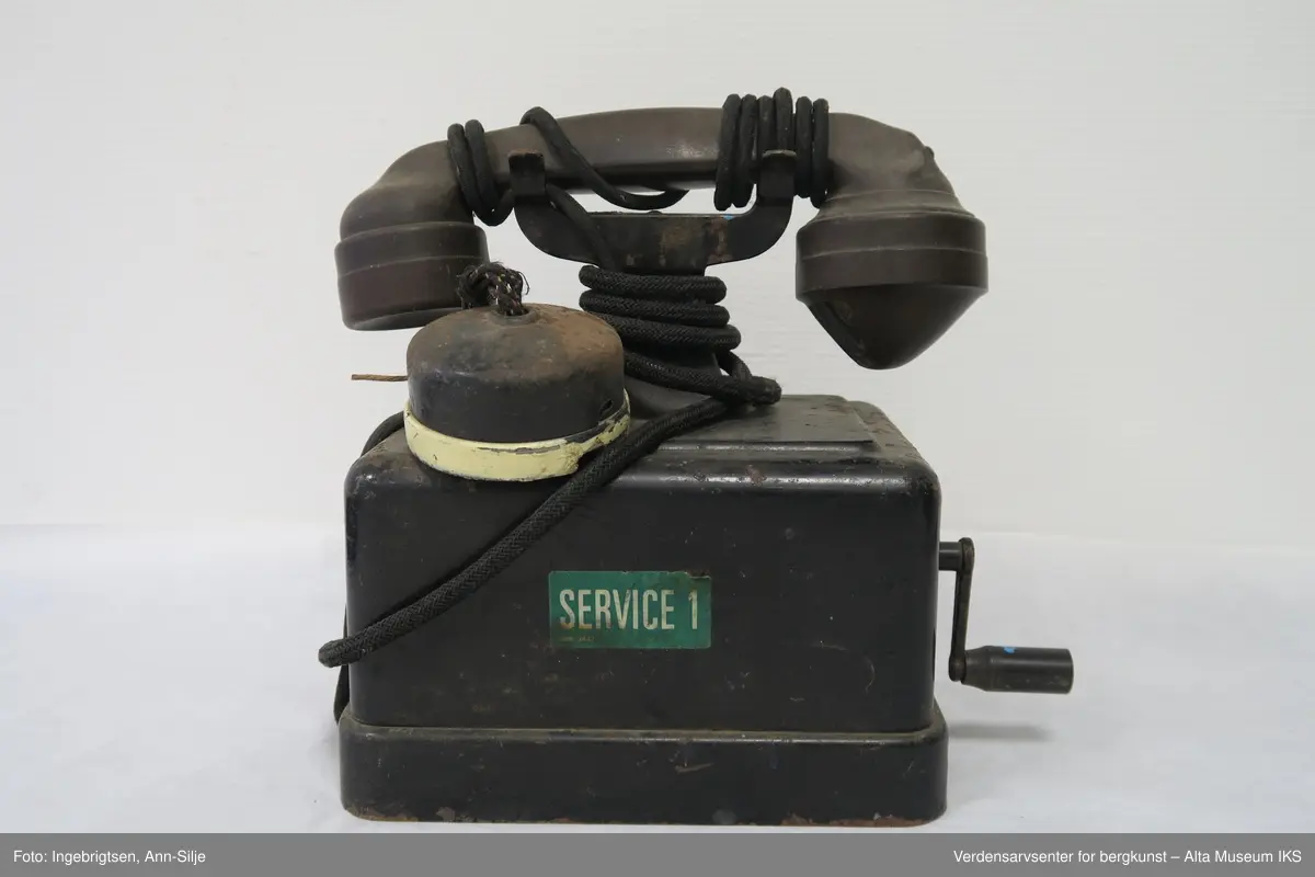 Prismeformet med bæreklo på toppen. Et telefonrør ligger på bærekloen. På boksen foran telefonrøret er det en ringeklokke. Plakett med tekst på fremsiden av boksen.