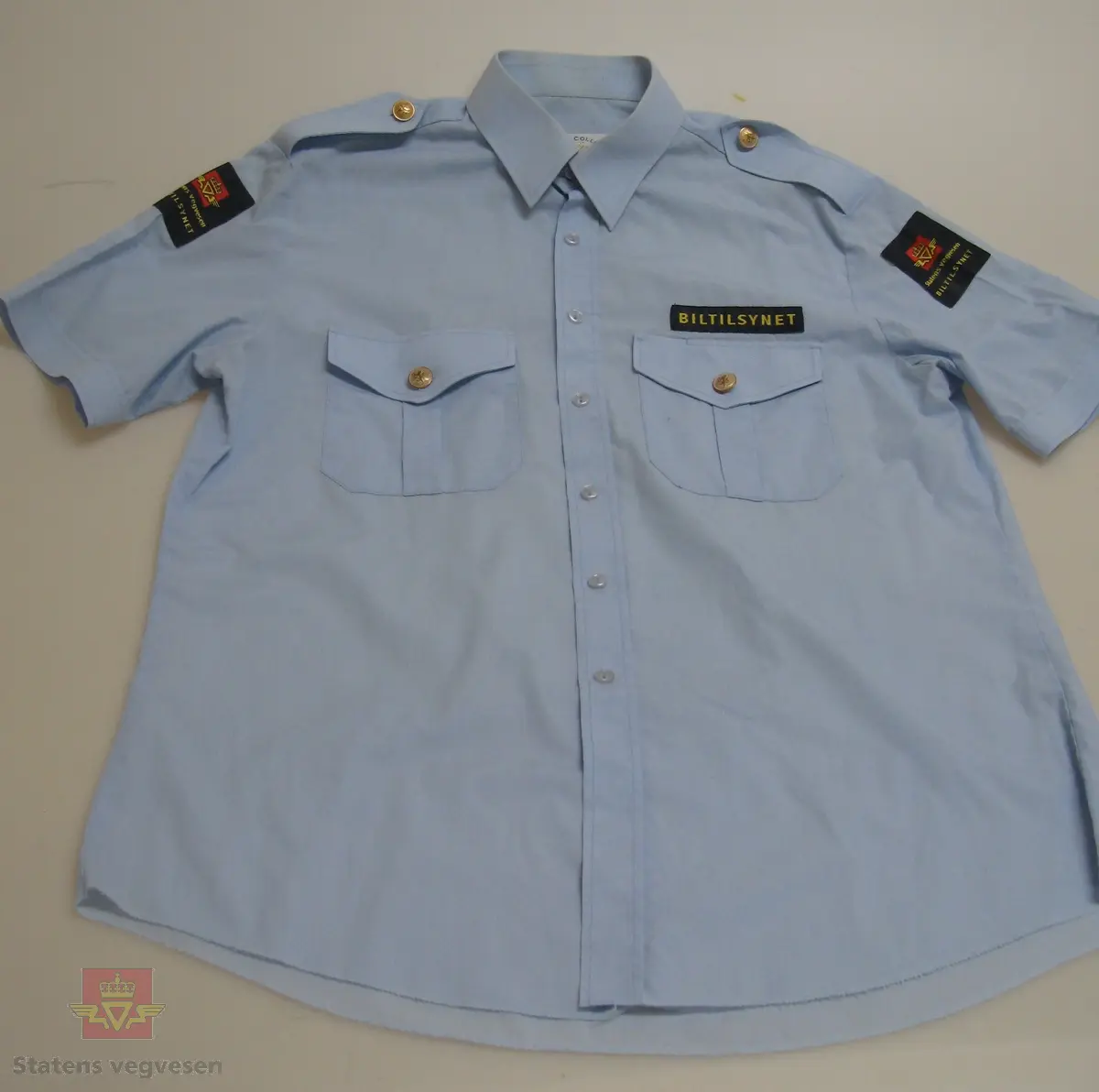 Kortermet lyseblå uniformskjorte i tynt bomullsstoff med skjortekrage, skulderklaffer og enkel knapping. Skjorta er i størrelse 41, herremodell. To brystlommer ,gullfargede knapper (type riksvåpen) både på skulderklaffer og lommer. Tøymerke med "BILTILSYNET" over venstre brystlomme. Påsydd firkantet merke med etatslogo og Biltilsynet, på både høyre og venstre arm.