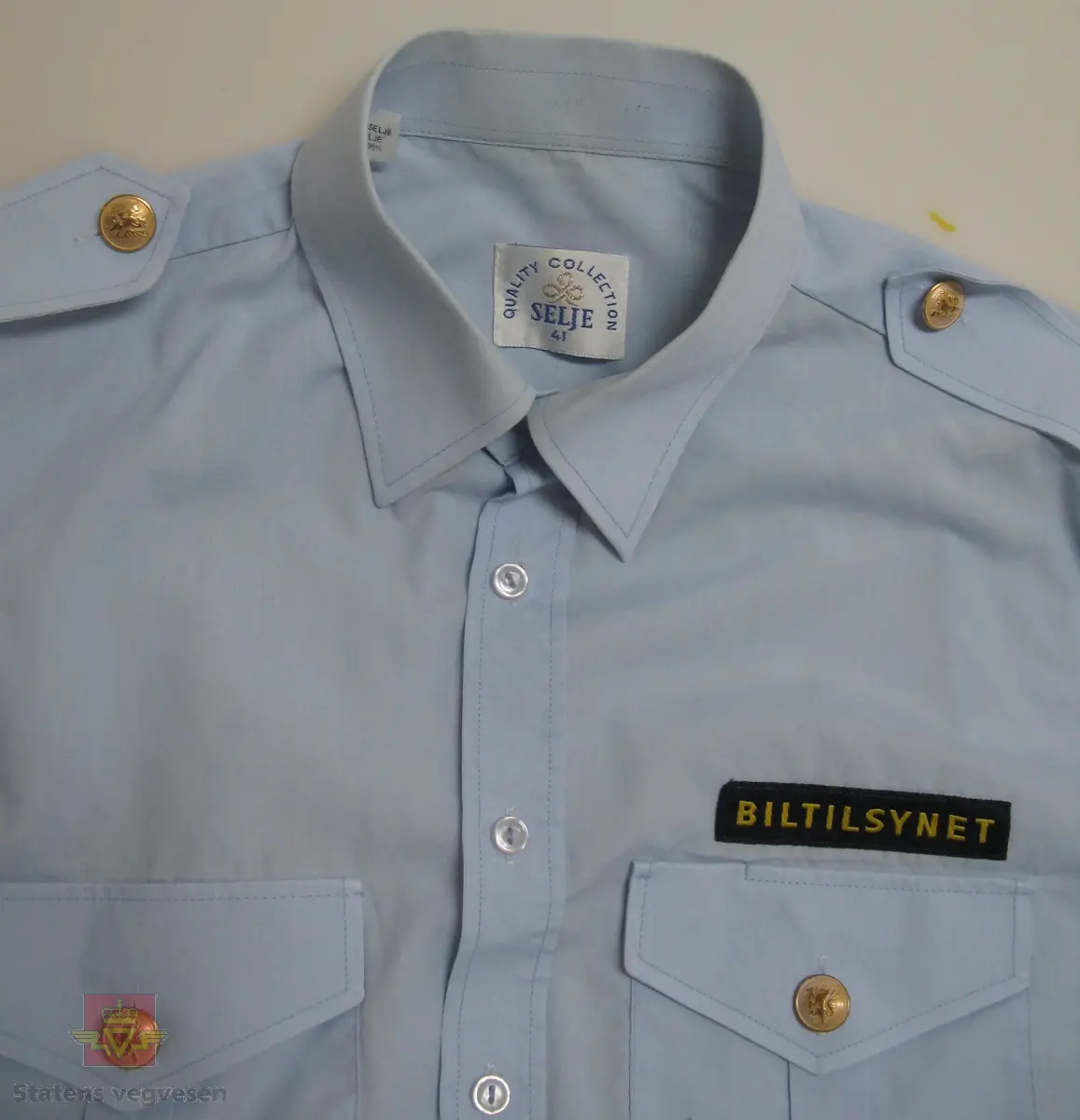 Kortermet lyseblå uniformskjorte i tynt bomullsstoff med skjortekrage, skulderklaffer og enkel knapping. Skjorta er i størrelse 41, herremodell. To brystlommer ,gullfargede knapper (type riksvåpen) både på skulderklaffer og lommer. Tøymerke med "BILTILSYNET" over venstre brystlomme. Påsydd firkantet merke med etatslogo og Biltilsynet, på både høyre og venstre arm.