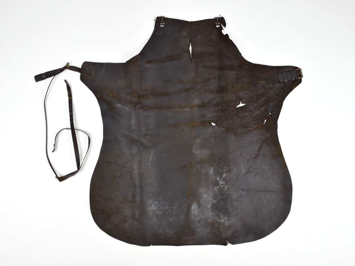 Smedförskinn av läder som är skuret i ett stycke. Skinnet har en rem runt halsen och en rem runt midjan. Spännena på remmarna är av metall.