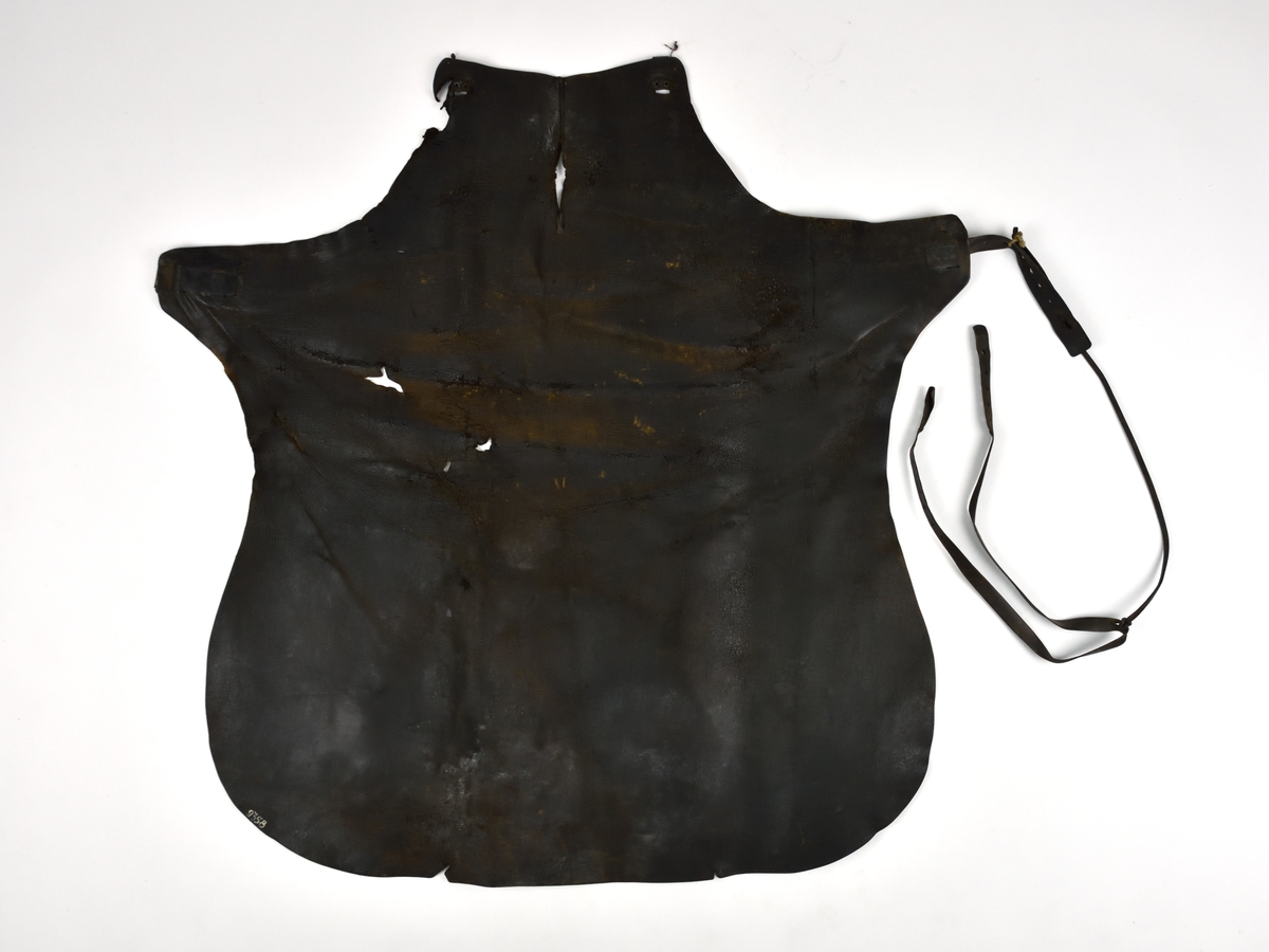 Smedförskinn av läder som är skuret i ett stycke. Skinnet har en rem runt halsen och en rem runt midjan. Spännena på remmarna är av metall.