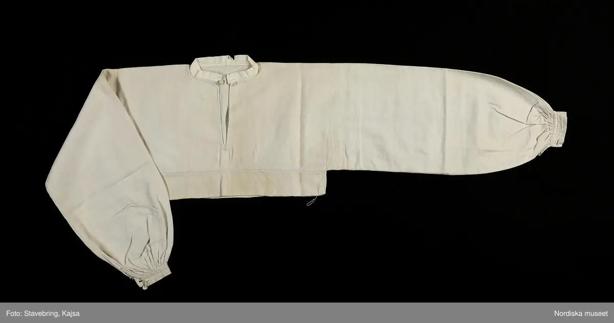 Huvudliggaren:
"Sorgdräkt för kvinna. Särk med 'hängseltåg', 'särkyvdel'; vit -'häsklä'; blått livstycke; svart tröja; huvudduk; 'svart näsduk'; hatt; 'raskförklä', 'sockar',  'hårbandsvifsa', huvudbindel."

Bilaga:
a: "8 Särk, övdel. av linne eller blågarn. med broderade linningar på överdelen"
c: "11. Hvit häsklä."
Anm: Kung Kerstin Larsdotters resa i Floda sn Dalarna 24/2 - 8/3 1912. Innehåller förteckning på inköp av dräktdelar från Floda, inv nr 117 216 - 117 239. Här finns  tillverkningstid och beskrivningar på vissa av dräktdelarna.  I vissa fall nämns också utförligt på vilka helgdagar dräktdelen  användes.

Katalogkort:
a: Hängselsärk
b. Överdel av halvlinne vävt i kypert.
c.  Halskäde av småmönstrat bomullstyg, liknande kambris, vävt  i kypertvariation med drällmönster.
d. Livstycke av mörkblå kraftig vadmal,
e. Tröja av grov svart vadmal

i. 
 Mönstervävt band med upplockat mönster i rött z-tvinnat ullgarn på vit linnebotten, med hjärtan, kryss och rombformer, 2 band om vardera 124 cm har vikts och sytts ihop så att det ena bildar en ögla och det andra har fria ändar avslutade med en tofs i bandets garn.
Läs mer om dräktskicket i Floda i: Albert Alm, Dräktalmanacka för Floda socken, Nordiska museet, Sthlm 1930
/Berit Eldvik 2005-04-14