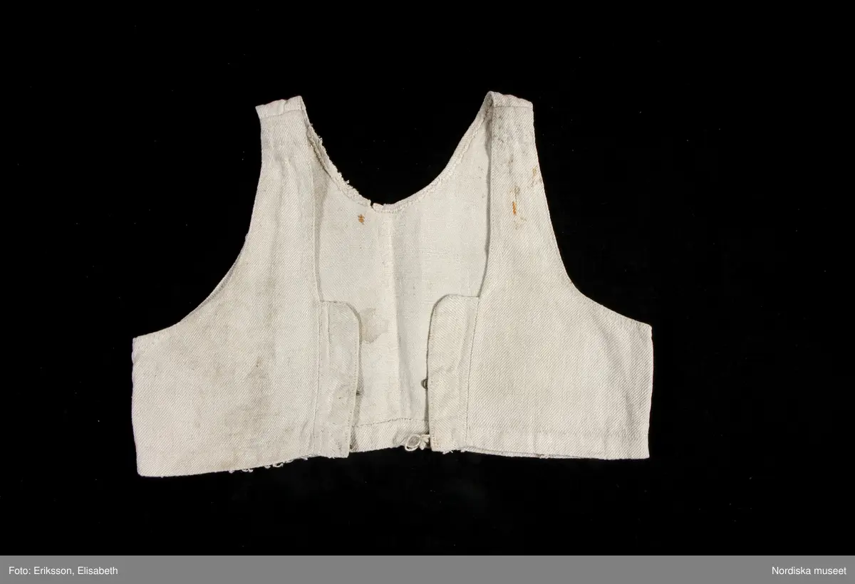 Livstycke, "bod" som varit fastsydd på en linnekjol. Av handspunnet lingarn, blångarn, i treskaftad kypert, "treskäft".  2 raka framstycken och ett ryggstycke, skarvat på axlarna. 3 påsydda band i ryggen som sömmarkeringar, blå med plockat mönster i vitt lingarn. Öppet framtill med 3 cm breda knäppkanter på båda sidorna med 2 par hakar och hyskor av mässing. Fåll nedtill med stygnrester av lingarn där den varit fastsydd mot kjolen.
Anm. sliten med fläckar och ett hål i ryggen.
Denna typ av livkjol, kallas i Mora och på Sollerön för vitsärk och användes sommartid. De kunde vävas mer eller mindre grova, och syddes mer eller mindre fint, släta, grovrynkade eller finrynkade och med grövre eller finare liv (bod) alltefter som man avsåg dem till grövsta vardagsbruk eller till helgdagsplagg.
Till fint hade de ofta liv av blommigt tyg men även röda eller gröna vadmalsliv förekom, de enklare hade vitt linneliv med bandgarnering. 
Vitsärkarna kom rätt tidigt ur bruk, sist var det de gamla gummorna, som nyttjade dem. Få är bevarade, ty det var vanligt att kvinnorna sveptes i dem.
Se Ella Odstedt: Folkdräkter i övre Dalarne sid 194-195.

Jfr. hel livkjol från Sollerön inv.nr 211804.
/Berit Eldvik 2007-09-21