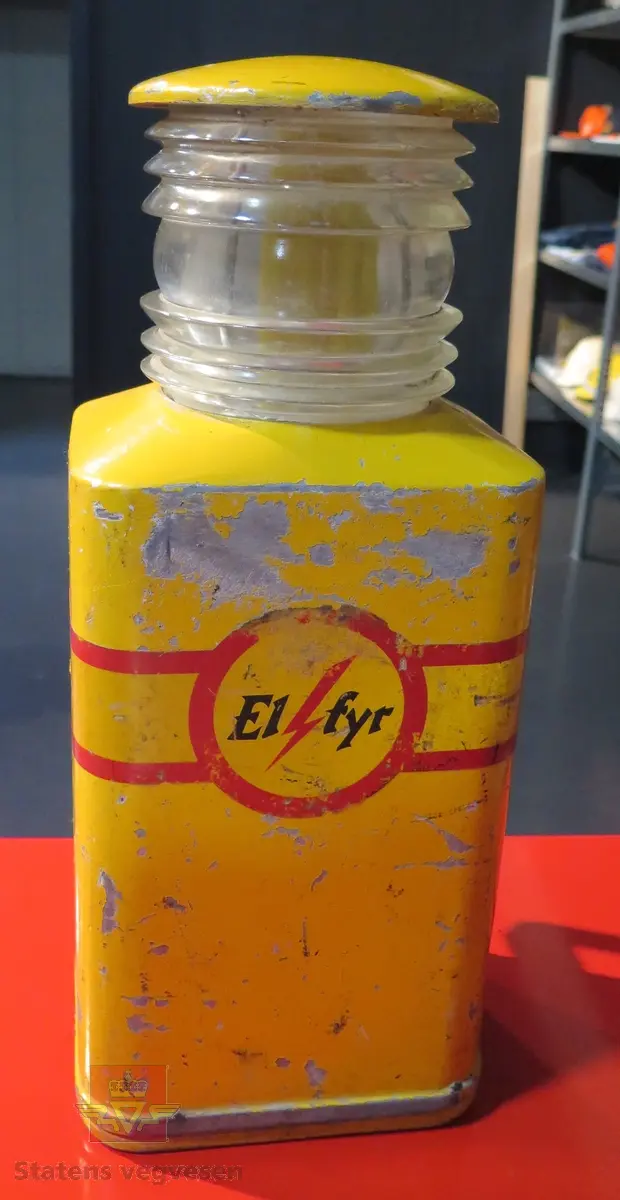 Rektangulær varsellykt (lampe). Den er gul med rødes striper. Drevet av fire Hellesens 1,5 volt batterier, seriekoblet til 6 volt, (her fjernet). Tekst "El-fyr".