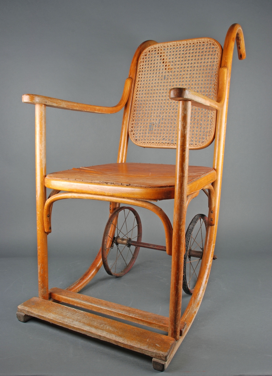 Rullestol i formpresset tre. Fotplate. Håndtak i forlengelsen av ryggen. Rotting rygg. Finert sete erstatter rotting. 2 hjul.