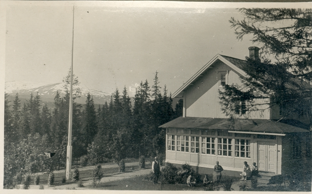 Gruppe utendørs. En gruppe menn, kvinner og barn sitter utenfor trehus med glassveranda. Flaggstang og opparbeidet hage. Granskog. Fjell i bakgrunnen.