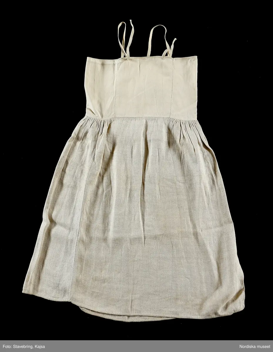 Huvudliggaren:
"Sorgdräkt för kvinna. Särk med 'hängseltåg', 'särkyvdel'; vit -'häsklä'; blått livstycke; svart tröja; huvudduk; 'svart näsduk'; hatt; 'raskförklä', 'sockar',  'hårbandsvifsa', huvudbindel."

Bilaga:
a: "8 Särk, övdel. av linne eller blågarn. med broderade linningar på överdelen"
c: "11. Hvit häsklä."
Anm: Kung Kerstin Larsdotters resa i Floda sn Dalarna 24/2 - 8/3 1912. Innehåller förteckning på inköp av dräktdelar från Floda, inv nr 117 216 - 117 239. Här finns  tillverkningstid och beskrivningar på vissa av dräktdelarna.  I vissa fall nämns också utförligt på vilka helgdagar dräktdelen  användes.

Katalogkort:
a: Hängselsärk
b. Överdel av halvlinne vävt i kypert.
c.  Halskäde av småmönstrat bomullstyg, liknande kambris, vävt  i kypertvariation med drällmönster.
d. Livstycke av mörkblå kraftig vadmal,
e. Tröja av grov svart vadmal

i. 
 Mönstervävt band med upplockat mönster i rött z-tvinnat ullgarn på vit linnebotten, med hjärtan, kryss och rombformer, 2 band om vardera 124 cm har vikts och sytts ihop så att det ena bildar en ögla och det andra har fria ändar avslutade med en tofs i bandets garn.
Läs mer om dräktskicket i Floda i: Albert Alm, Dräktalmanacka för Floda socken, Nordiska museet, Sthlm 1930
/Berit Eldvik 2005-04-14