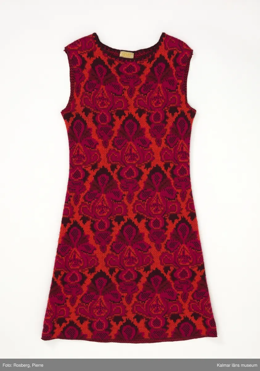 KLM 39302:13. Klänning. 1970-tal. Av jaquardstickad ylle, fyra färgs jaquard med köperbaksida. Mönster; växtdelar i stora rombformer i röda nyanser på orange botten. Knälång, rak modell. Rund halsringning, maskinlaskad med mörkrött garn. Ärmslös. Ärmhål och fåll sydda med overlocksöm. Nära fållen på avigsidan finns en fastsydd remsa av rött fodertyg, troligen rest från ett bortklippt helfoder. Fastsydd textileikett mitt bak med invävd text: MMT Design Katja of Sweden MILD 25 38.