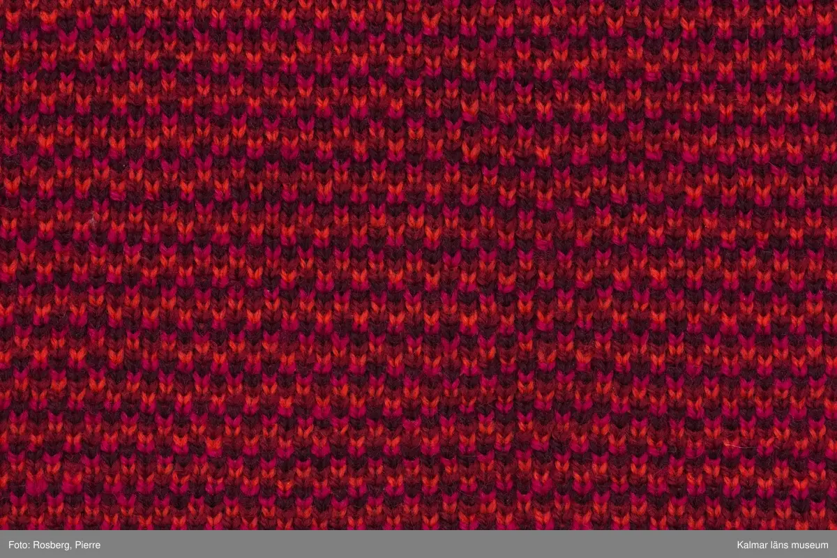 KLM 39302:13. Klänning. 1970-tal. Av jaquardstickad ylle, fyra färgs jaquard med köperbaksida. Mönster; växtdelar i stora rombformer i röda nyanser på orange botten. Knälång, rak modell. Rund halsringning, maskinlaskad med mörkrött garn. Ärmslös. Ärmhål och fåll sydda med overlocksöm. Nära fållen på avigsidan finns en fastsydd remsa av rött fodertyg, troligen rest från ett bortklippt helfoder. Fastsydd textileikett mitt bak med invävd text: MMT Design Katja of Sweden MILD 25 38.