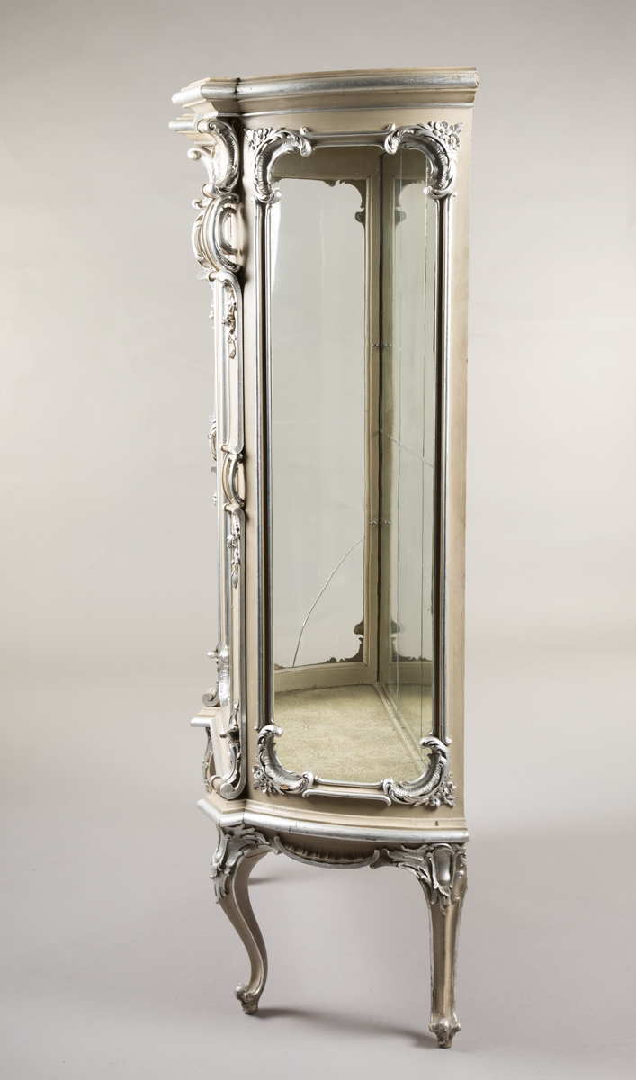 Vitrineskap i nyrokokko. Front og sider i glass, bakveggen av tre, på innsiden dekket av blyinnfattet speil. Sideveggene har buet glass. Alt treverk rikt utskåret. Malt lysegrått med forsølvete ornamenter. 3 hyller trukket med stoff, 1 dør. 4 cabriolebein med voluttavslutning. Øverste profilerte gesims med stort, utskåret toppstykke hvor to speil er innfelt.