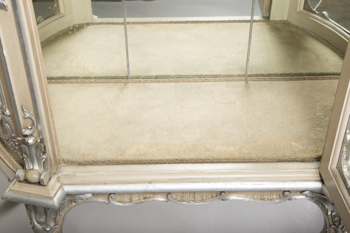 Vitrineskap i nyrokokko. Front og sider i glass, bakveggen av tre, på innsiden dekket av blyinnfattet speil. Sideveggene har buet glass. Alt treverk rikt utskåret. Malt lysegrått med forsølvete ornamenter. 3 hyller trukket med stoff, 1 dør. 4 cabriolebein med voluttavslutning. Øverste profilerte gesims med stort, utskåret toppstykke hvor to speil er innfelt.
